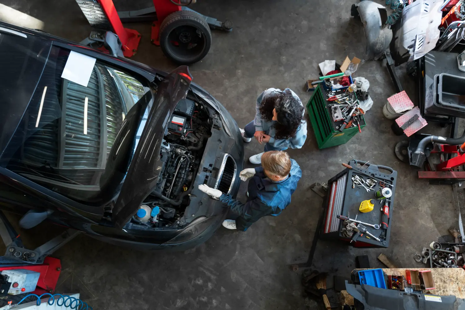 eine Draufsicht auf eine Autowerkstatt, in der ein Mechaniker einem Kunden etwas über einen Automotor erklärt